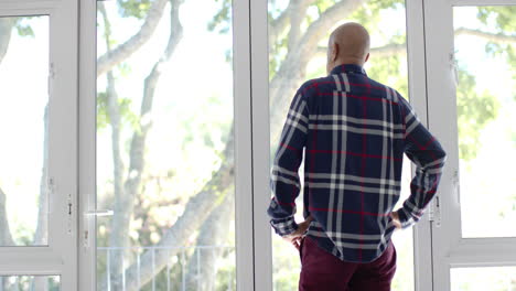 Thoughtful-senior-biracial-man-looking-through-window-at-home,-slow-motion