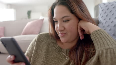 Happy-plus-size-biracial-woman-relaxing-in-living-room-using-smartphone,-slow-motion