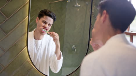 Biracial-man-brushing-teeth-in-morning,-looking-in-bathroom-mirror,-slow-motion