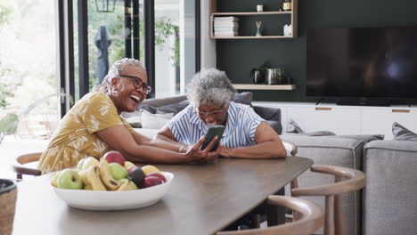 Senior-african-american-female-friends-using-smartphone-and-laughing-in-dining-room,-slow-motion