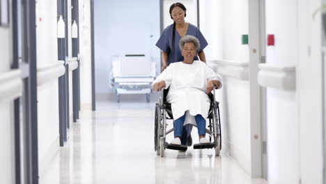 African-american-female-doctor-walking-with-senior-woman-in-wheelchair-in-hospital,-slow-motion