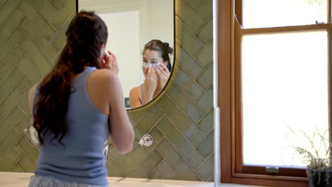 Biracial-teenage-girl-putting-on-under-eye-masks-looking-in-bathroom-mirror,-copy-space,-slow-motion