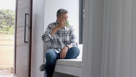 Relaxed-senior-biracial-man-sitting-at-window-and-drinking-coffee-at-home,-copy-space,-slow-motion