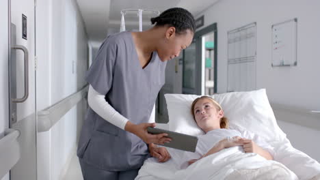 Diverse-female-doctor-and-girl-patient-lying-on-bed-using-tablet-in-hospital,-slow-motion