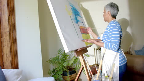 Focused-senior-biracial-woman-wearing-apron-and-painting-on-big-canvas-at-home,-slow-motion