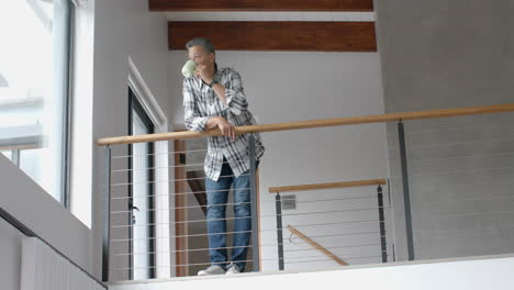 Senior-biracial-man-looking-out-of-window,-drinking-coffee-on-staircase,-copy-space,-slow-motion