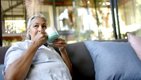 Biracial-senior-woman-sitting-on-couch,-drinking-tea-and-looking-side-at-home