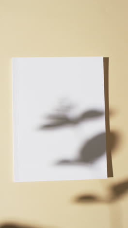 Vertical-video-of-shadow-of-plant-over-book-with-blank-pages-and-copy-space-on-yellow-background