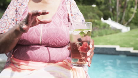 Glückliche-ältere-Afroamerikanische-Frau-Mit-Sonnenbrille-Am-Pool,-Die-Cocktails-Trinkt,-Zeitlupe