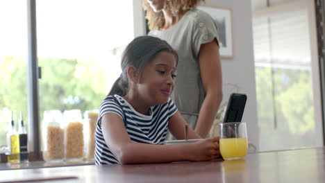 Happy-biracial-daughter-using-smartphone-in-sunny-kitchen-with-mother,-slow-motion