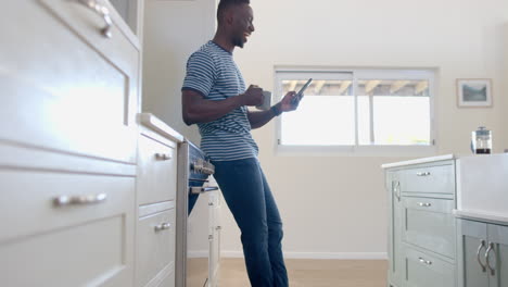 Happy-african-american-man-drinking-coffee-and-using-smartphone-in-sunny-kitchen,-slow-motion