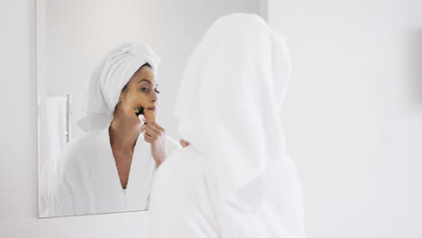 Happy-biracial-woman-applying-beauty-face-mask-looking-in-mirror-in-bathroom,-slow-motion