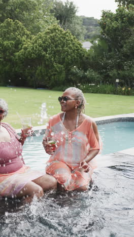 Vertical-video-of-senior-african-american-female-friends-sitting-in-pool-with-drinks,-slow-motion