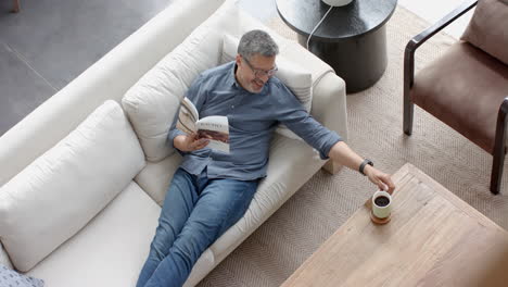 Glücklicher-älterer-Mann-Mit-Gemischter-Abstammung-Auf-Der-Couch,-Der-Kaffee-Trinkt-Und-Im-Wohnzimmer-Ein-Buch-Liest,-Zeitlupe