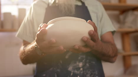 Mid-section-of-biracial-potter-with-long-beard-holding-bowl-in-pottery-studio,-slow-motion