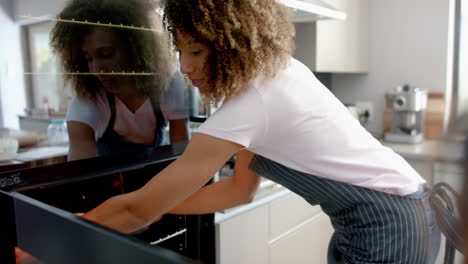 Happy-biracial-mother-putting-cakes-in-oven,-baking-with-daughter-in-kitchen,-slow-motion