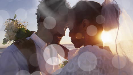 Animation-of-light-spots-over-happy-african-american-bride-and-groom-embracing-on-beach-at-wedding