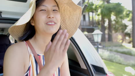 Glückliche-Asiatische-Frau-Mit-Sonnenhut,-Die-Im-Kofferraum-Eines-Autos-Sitzt-Und-An-Einem-Sonnigen-Tag-Einen-Kuss-Zuwirft,-Zeitlupe