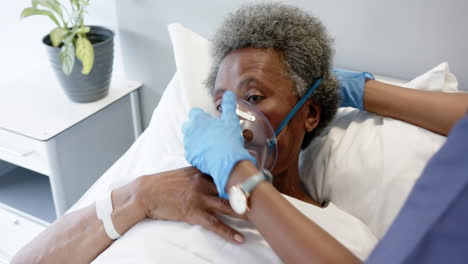 African-american-female-doctor-applying-oxygen-mask-to-senior-woman-in-hospital-room,-slow-motion