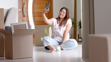 Happy-plus-size-biracial-woman-taking-selfie-sitting-on-floor-of-new-home,-copy-space,-slow-motion