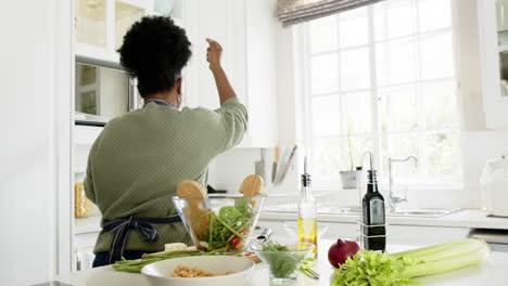 Happy-african-american-senior-woman-dancing-with-smartphone-and-headphones-in-kitchen,-slow-motion