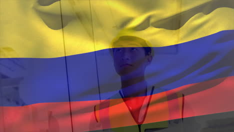 Animation-of-flag-of-colombia-waving-over-smiling-asian-male-architect-in-hard-hat-holding-blueprint