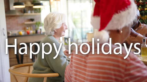 Animation-of-happy-holidays-text-over-diverse-senior-friends-sitting-at-table-at-christmas