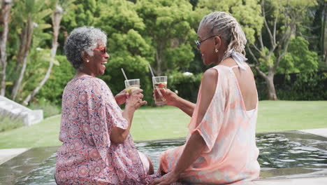 Happy-senior-african-american-female-friends-making-a-toast-with-drinks-sitting-by-pool,-slow-motion