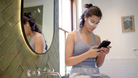 Biracial-teenage-girl-wearing-under-eye-masks-using-smartphone-in-bathroom,-copy-space,-slow-motion