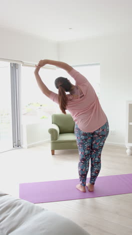 Vertical-video-of-plus-size-biracial-woman-standing-practicing-yoga,-stretching-at-home,-slow-motion