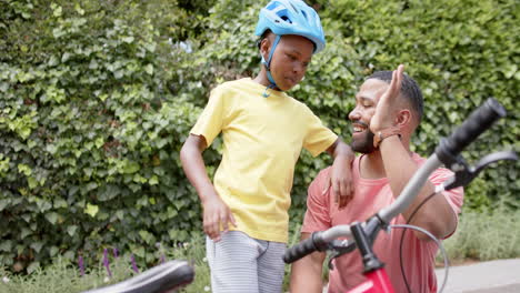 Feliz-Padre-E-Hijo-Afroamericanos-Con-Bicicleta-Chocando-Los-Cinco-En-El-Jardín,-Cámara-Lenta