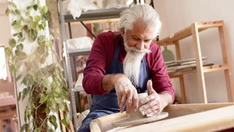 Focused-senior-biracial-potter-with-long-beard-using-potter's-wheel-in-pottery-studio,-slow-motion