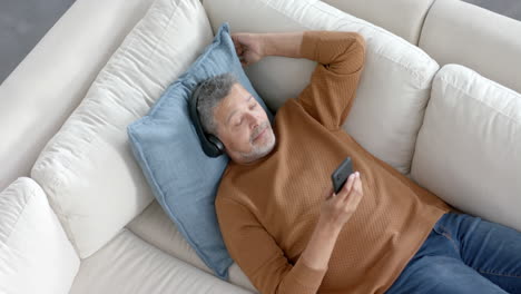 Senior-biracial-man-on-couch-in-headphones-listening-to-music-on-smartphone,-copy-space,-slow-motion