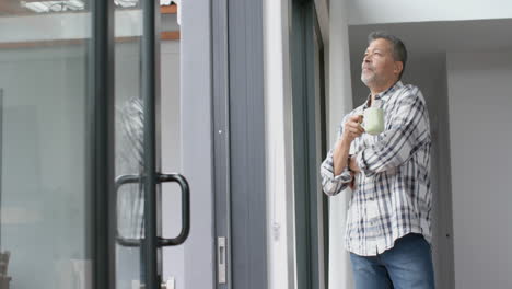 Thoughtful-senior-biracial-man-with-coffee-looking-out-of-window-at-home,-copy-space,-slow-motion