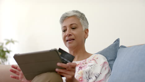 Glückliche-ältere-Frau-Mit-Gemischter-Abstammung-Auf-Der-Couch,-Die-Eine-Kreditkarte-Hält-Und-Zu-Hause-Ein-Tablet-Benutzt,-Zeitlupe