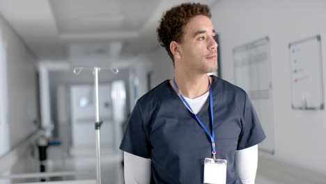 Portrait-of-happy-biracial-male-doctor-wearing-scrubs-in-hospital-corridor,-copy-space,-slow-motion