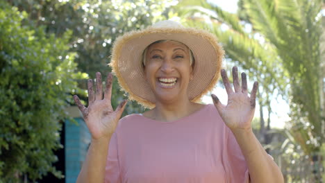 Portrait-of-happy-senior-biracial-woman-with-dirty-hands-wearing-hat-in-garden-at-home,-slow-motion