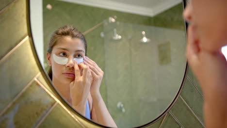 Focused-biracial-teenage-girl-putting-on-under-eye-masks-looking-in-bathroom-mirror,-slow-motion