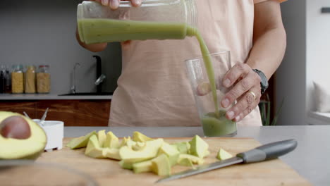 Midsection-of-senior-biracial-man-pouring-healthy-smoothie-into-glass-in-kitchen,-slow-motion