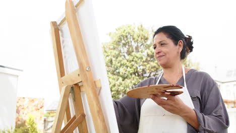 Happy-biracial-senior-woman-painting-picture-and-smiling-in-sunny-garden,-slow-motion,-copy-space