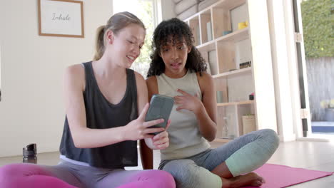 Happy-diverse-women-sitting-on-yoga-mats-and-using-smartphone,-slow-motion