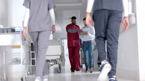 Diverse-male-doctors-discussing-work-using-tablet-walking-in-hospital-corridor,-slow-motion