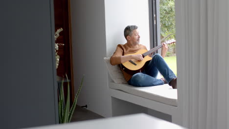 Focused-senior-biracial-man-sitting-on-window-seat-playing-guitar-at-home,-copy-space,-slow-motion