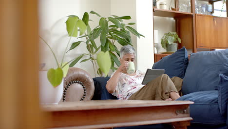 Glückliche-ältere-Frau-Mit-Gemischter-Abstammung,-Die-Auf-Der-Couch-Sitzt,-Tee-Trinkt-Und-Zu-Hause-Ein-Tablet-Benutzt,-Zeitlupe