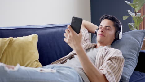 Happy-biracial-man-in-headphones-lying-on-couch-using-smartphone-and-closing-eyes,-slow-motion