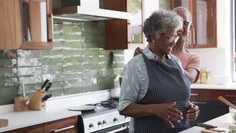 Happy-senior-african-american-female-friends-putting-on-apron-in-kitchen,-copy-space,-slow-motion