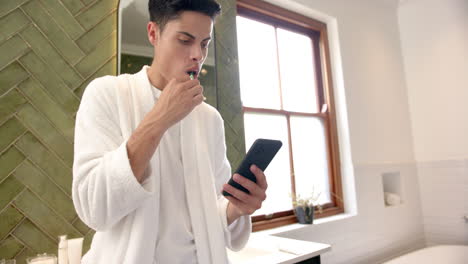 Biracial-man-brushing-teeth-and-using-smartphone-in-bathroom-in-the-morning,-copy-space,-slow-motion