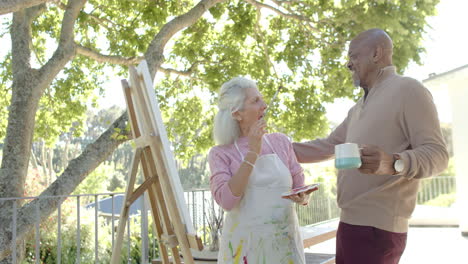 Happy-senior-biracial-couple-painting-and-drinking-coffee-at-balcony-at-home,-slow-motion