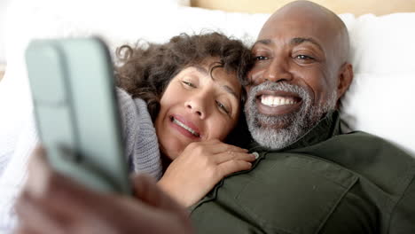 Happy-mature-diverse-couple-lying-on-bed-using-smartphone-at-home,-slow-motion