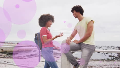Animation-of-purple-spots-over-happy-biracial-couple-talking-on-beach-promenade